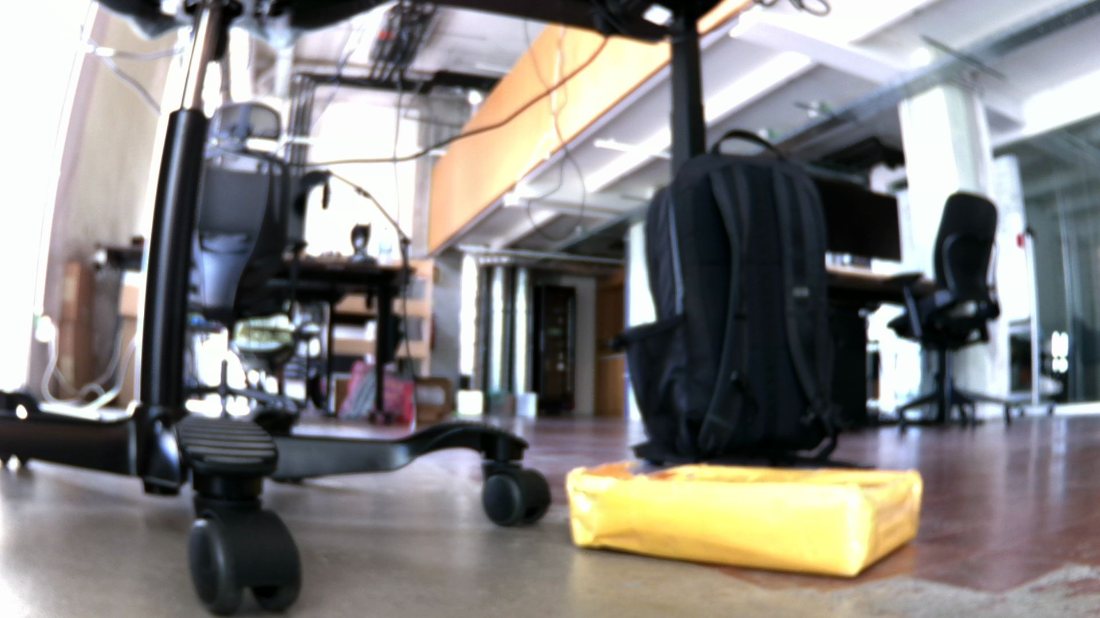 Robot camera frame: a yellow padded mailer on the floor beside a chair base, at touching distance